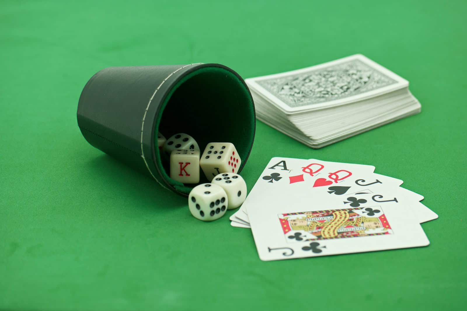 Dice spilling from a cup, playing cards, and a stack of cards on a green gaming table.